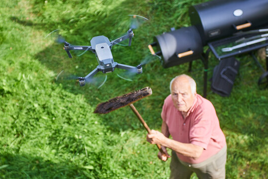 Old senior man upset by a flying drone over his garden. The concept of spying on neighbors and their privacy. Man trying to knock down a drone from the sky over his own garden with a broom. Above view