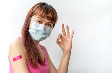 Asian woman wear face mask using adhesive bandage with pink at arm and showing O.K. gesture after injection vaccine. Covid 19 vaccine disease preparing.