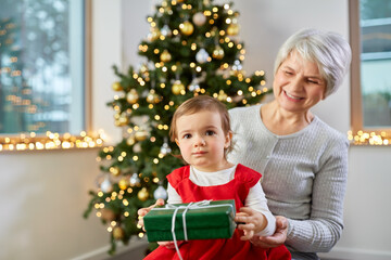 Obraz na płótnie Canvas christmas, holidays and family concept - happy grandmother and baby granddaughter with gift box at home