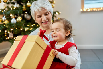 Obraz na płótnie Canvas christmas, holidays and family concept - happy grandmother and baby granddaughter opening gift box at home