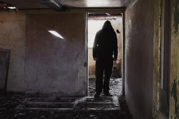 A hooded man standing in the doorway of a ruined, abandoned house