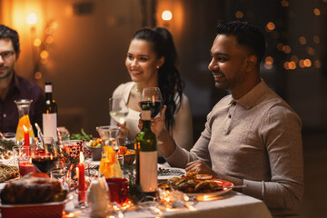 holidays, party and celebration concept - multiethnic group of happy friends having christmas dinner at home and drinking wine