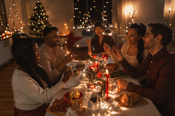 holidays and celebration concept - multiethnic group of happy friends with sparklers having christmas dinner at home