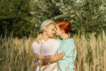 Lesbian young woman and girlfriend hug standing in high dry field grass against green trees in summer park at bright sunlight