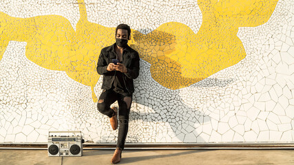 Young Afro man using mobile smartphone while standing against urban wall and listening music with headphones and vintage boombox