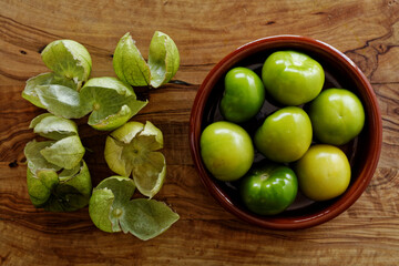 Husked tomatillos in clay bowl along with their husks lying on cutting board