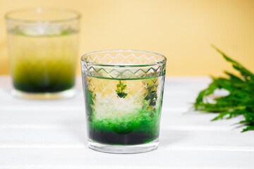 Green Chlorophyll or Chlorella drink in glass with water on white table, Orange Background. Green Drops in clear water, Antioxidant food and drinks.