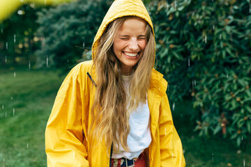 Playful young blonde woman smiling wearing yellow raincoat during the rain in the park. Cheerful...
