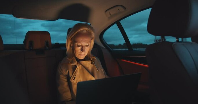 Mature businesswoman working on laptop while sitting on backseat in car late in evening