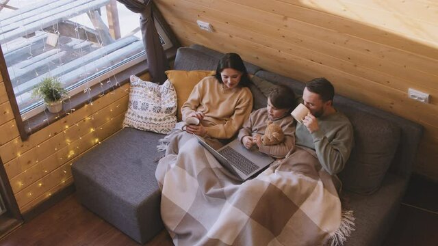 High angle slowmo of father, mother and daughter sitting together on couch with their legs wrapped in blanket. They are watching movie on laptop and drinking hot tea in their cozy cabin on winter day