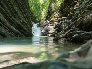 A beautiful transparent waterfall in layered rocks quickly flows into an aquamarine font. Stones in the foreground. Mountain landscape