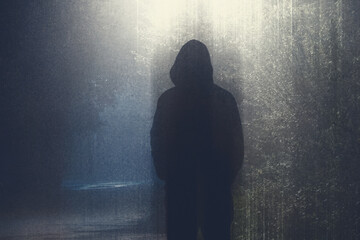 A hooded figure silhouetted against a street light lighting up a country road. On a foggy night. With a grunge edit