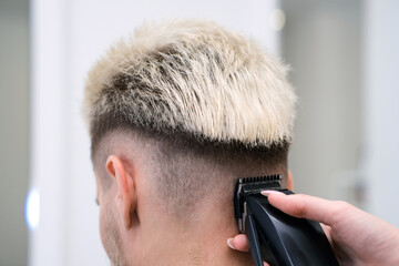 haircut process of blond young man with hair clipper in armchair in barbershop salon