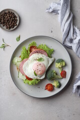 beautiful healthy food healthy breakfast sandwich with poached egg,lettuce, broccoli and cherry tomatoes on a plate on a gray background under concrete top view, with a knife and a napkin modern style