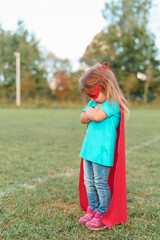 little girl in superhero costume with red cape is standing and taking offense on lawn in fresh air. child rebels and does not accept rules of game, gets upset with them for losing or being punished.