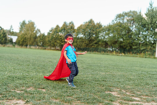 little boy in a superhero costume with a red cape runs across the lawn in the fresh air. The child fantasizes and pretends to be a superhero. A place for text or advertising. Copy space