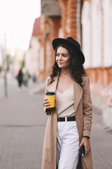 A beautiful brunette girl in a fashionable trench coat and hat walks through the streets of the city and drinks hot coffee in the autumn outdoors