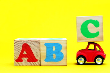 English letters ABC on wooden blocks on a yellow background. A toy car carrying a block with the letter C on the roof.