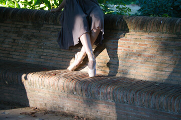 classical ballet dancer sitting on a bench in a park at the bottom of trees. The dancer has her feet in dancing position. Classical ballet concept