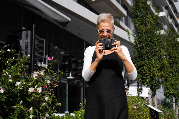 A beautiful senior woman is walking the streets of a city she is visiting and taking photos with a photo camera.