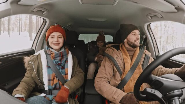 Shot From Inside Of Moving Car Happy Man Driving And Chatting With His Wife And Cute 8-year-old Girl Sitting In Passenger Seat And Backseat While Traveling In Forest Area In Winter