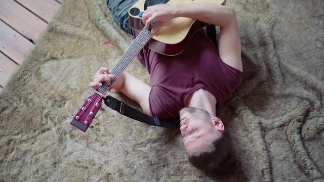 Man with acoustic guitar singing and playing guitar in lying position. Top view. Escape from boredom. Handsome caucasian male guitar player practice lying floor Creative man bored composes a song