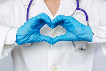 Cropped shot of a young female doctor in a white coat with a stethoscope around her neck forming a heart shape with her hands in blue gloves isolated on a gray background