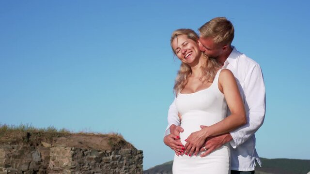 Tenderness in couple in love. A man is embracing his pregnant wife, rubbing her belly and kissing her neck and cheek against th blue sky in the morning 