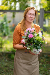 Gardening, floristics and middle aged people concept - portrait of woman in beige garden apron with bunch of flower over garden background.