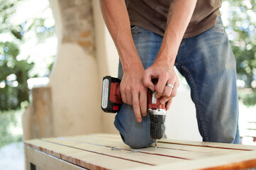 Skilled young male worker is using power screwdriver drilling during construction wooden bench, do it yourself