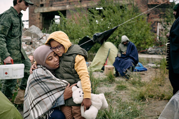 Sad Arabian woman hugging daughter with toy while army medic moving with first aid box behind them...