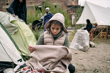 Sad middle-eastern refugee girl in hooded vest holding plaid while freezing in tent camp for migrants - obrazy, fototapety, plakaty