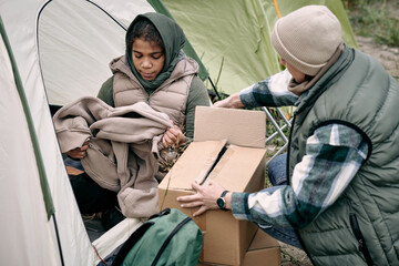 Middle-eastern refugee family in warm clothes unpacking boxes while getting settled into tent after...