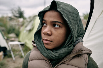 Sad Black refugee girl with covered head looking up hopefully, tent camp for migrant behind her
