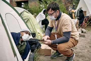 Young social worker with badge crouching at tent and making notes in tablet while talking to refugee - obrazy, fototapety, plakaty