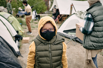Portrait of mixed race child in hoodie, vest, eyeglasses and cloth mask standing in refugee camp