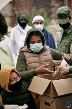 PHand of social worker putting products into cardboard box to refugee children in masks under control of army soldier