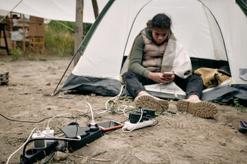 Middle-eastern migrant girl sitting in tent at socket outlets and using smartphone while charging...