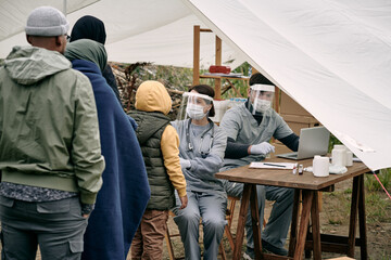 Medical specialists in protective masks sitting at table in tent and checking health of migrants...