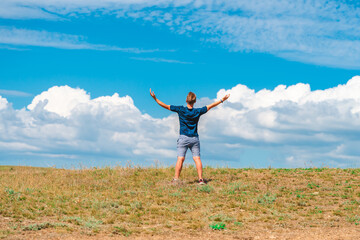 Rear view of a young man standing with his arms open against the background of a blue sky with clouds. The concept of freedom - Powered by Adobe