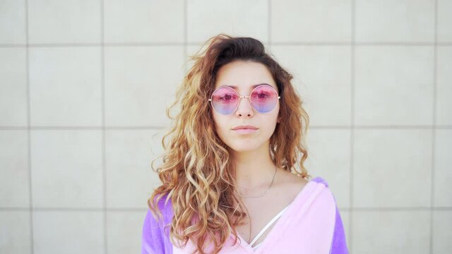 Close up portrait of a young pretty woman in pink and purple round glasses and curly red hair looking at the camera. Attractive caucasian hipster girl in sunglasses on urban wall background. Outdoors