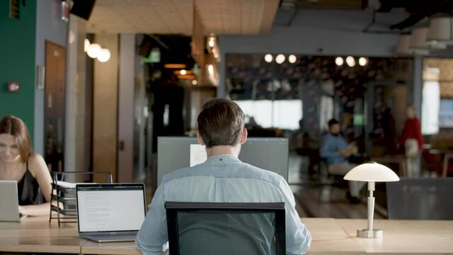 Unrecognizable businessman sitting at the big monitor at his workplace. Back view of manager looking at computer screen in office or co-working with his colleagues at background. Occupation concept