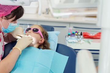 little girl at dentist's appointment at children's dental clinic. child is sitting with his mouth wide open, in examination chair with darkening glasses. professional dentist examines children's teeth