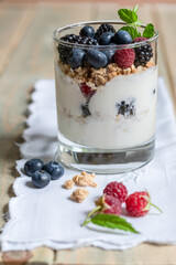 Yogurt with granola, fresh blueberries, blackberries and raspberries in a glass on an old wooden background. front view, close up, select focus.