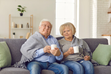 Happy mature couple sitting together on sofa under blanket and holding hot drinks in cups in hands at home with room interior at background. Elderly people happy lifestyle, family happiness concept