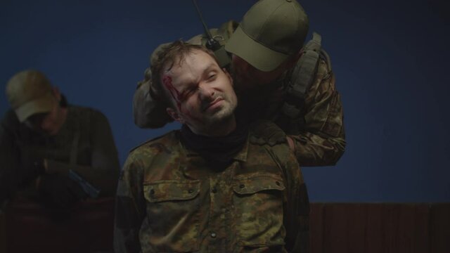 Portrait of injured prisoner of war in camouflage during interrogation indoors. Brutal male soldier intimidating enemy, putting gun to captive head, getting information from combatant