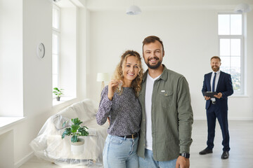 Happy young married couple looking at camera, smiling and showing key to new home standing in empty...
