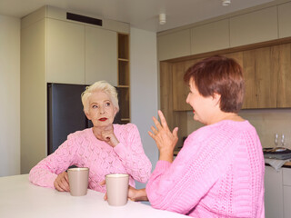 two stylish senior women in pink sweaters sitting with mugs at modern kitchen gossiping. Friendship, talk, gossip, relationships, news, family concept  