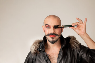 Portrait of drag queen looking at camera while covering one eye with makeup brush. Man with beard and mustache
