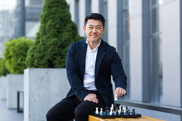 Cheerful Asian businessman playing chess in the yard smiling and looking forward to the camera